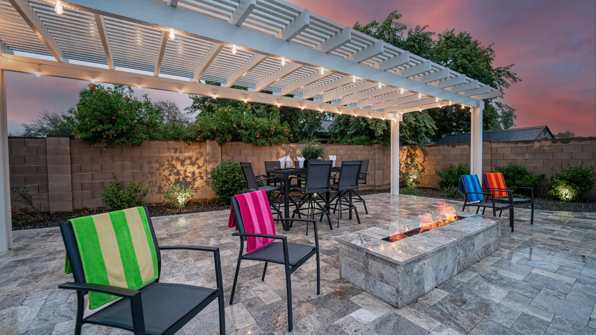 The image depicts a cozy fire pit area surrounded by colorful seating, with an outdoor dining table placed under a pergola. The setting is welcoming, perfect for memorable summer barbecues or intimate evening conversations by the fire.