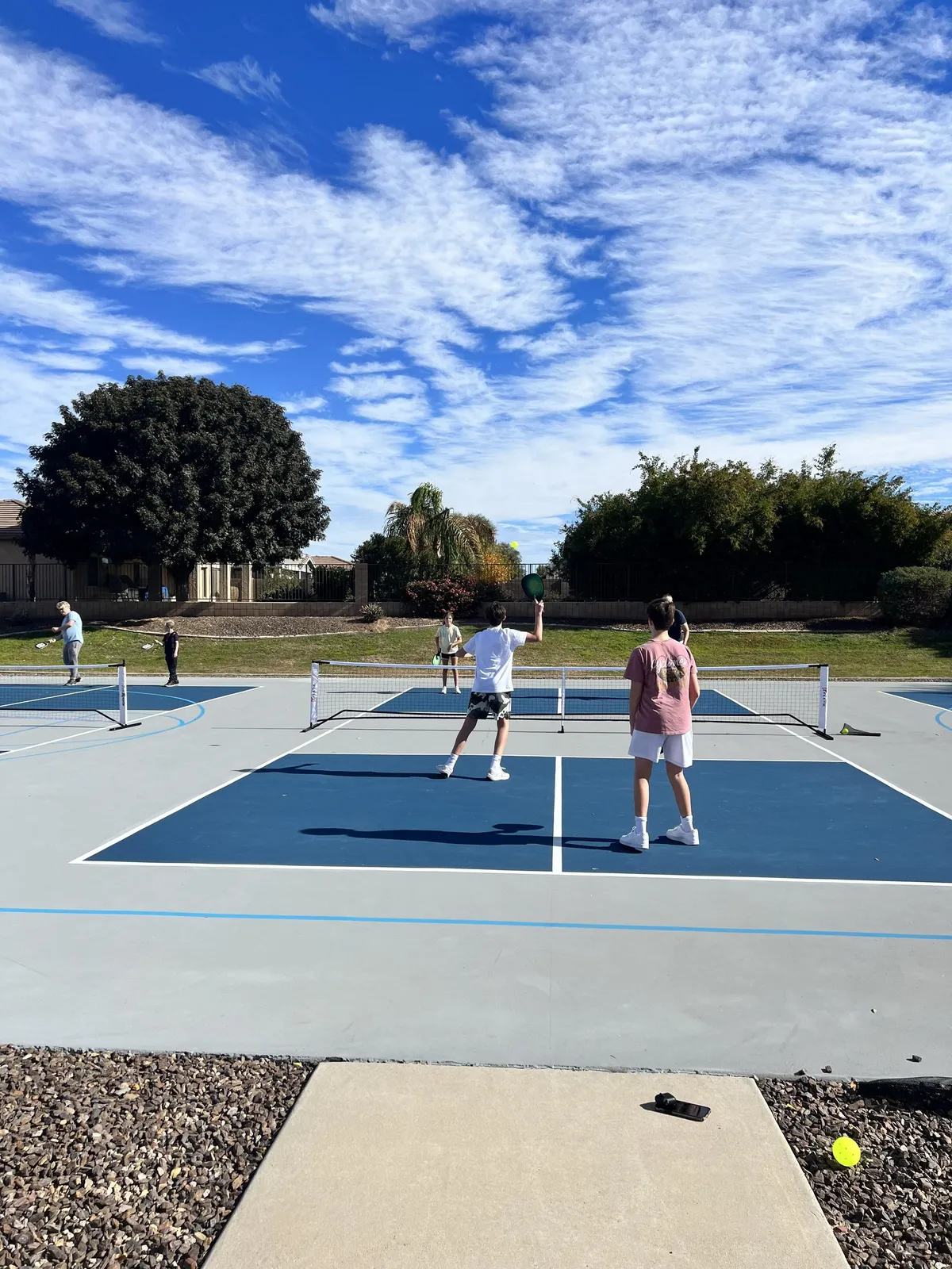 The image shows tennis players on a court, actively engaging in a game. The scene is set outdoors with greenery in the background, giving a sense of a community park area. The tennis net and the players' dynamic posture capture the spirit of outdoor recreation and friendly competition.
