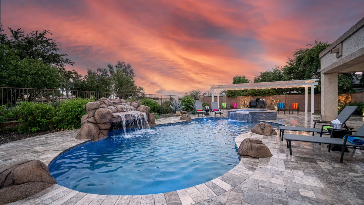 A sparkling swimming pool with a waterfall feature, colorful chairs for lounging, and an expansive sunset sky. There’s a sense of celebration and relaxation, with friends or family gathering to enjoy the inviting outdoor space.