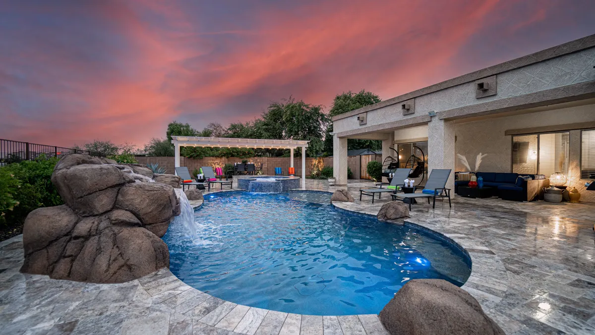This image features a swimming pool surrounded by an outdoor seating area, a waterfall feature, and a dramatic sunset sky. The atmosphere is serene yet vibrant, highlighting the abundance of outdoor amenities and space for relaxation or socializing.
