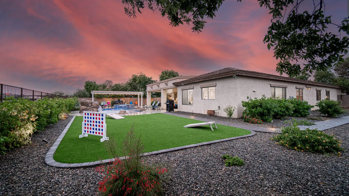 The scene features a lush green lawn set up for colorful outdoor games, with a view of the swimming pool and a dining area shaded under a pergola. The landscaping is vibrant, and the whole space is designed for playful family gatherings and relaxed outdoor living.