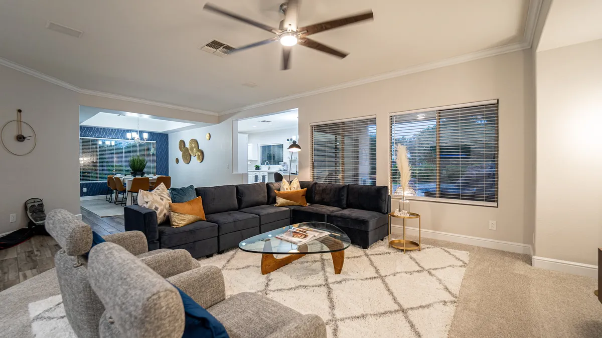 A spacious, sun-drenched living room featuring a large sectional sofa with decorative pillows, a sleek glass coffee table, and expansive windows that invite natural light. The room exudes a cozy and welcoming atmosphere, ideal for social gatherings or family movie nights.