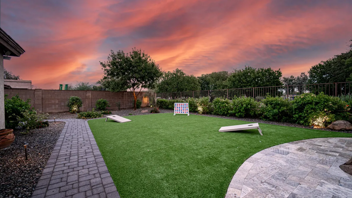 An inviting artificial lawn at sunset with a lively game of cornhole set up in the center, surrounded by lush greenery. The golden sky creates a playful and relaxed atmosphere, perfect for outdoor games and gatherings.