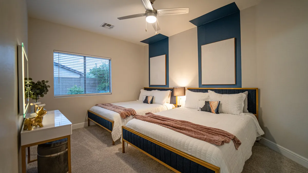 A bright and cozy guest room featuring two beds with decorative pillows, a stylish bedside lamp, and a window looking out to lush greenery. The room exudes warmth and flexibility for different guest arrangements.