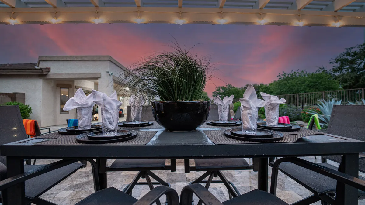 An elegantly set outdoor dining table with decorative plant centerpieces and colorful accents on the chairs. The area is prepared for an evening meal, suggesting an inviting atmosphere for al fresco dining under the stars.