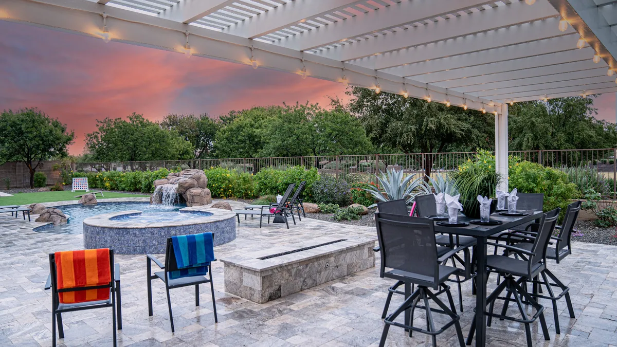 An inviting outdoor scene featuring a hot tub, a spacious dining table set for gatherings, a waterfall feature, and a landscaped garden under a glowing sunset sky. The area is set up for socializing, relaxing, and enjoying nature.