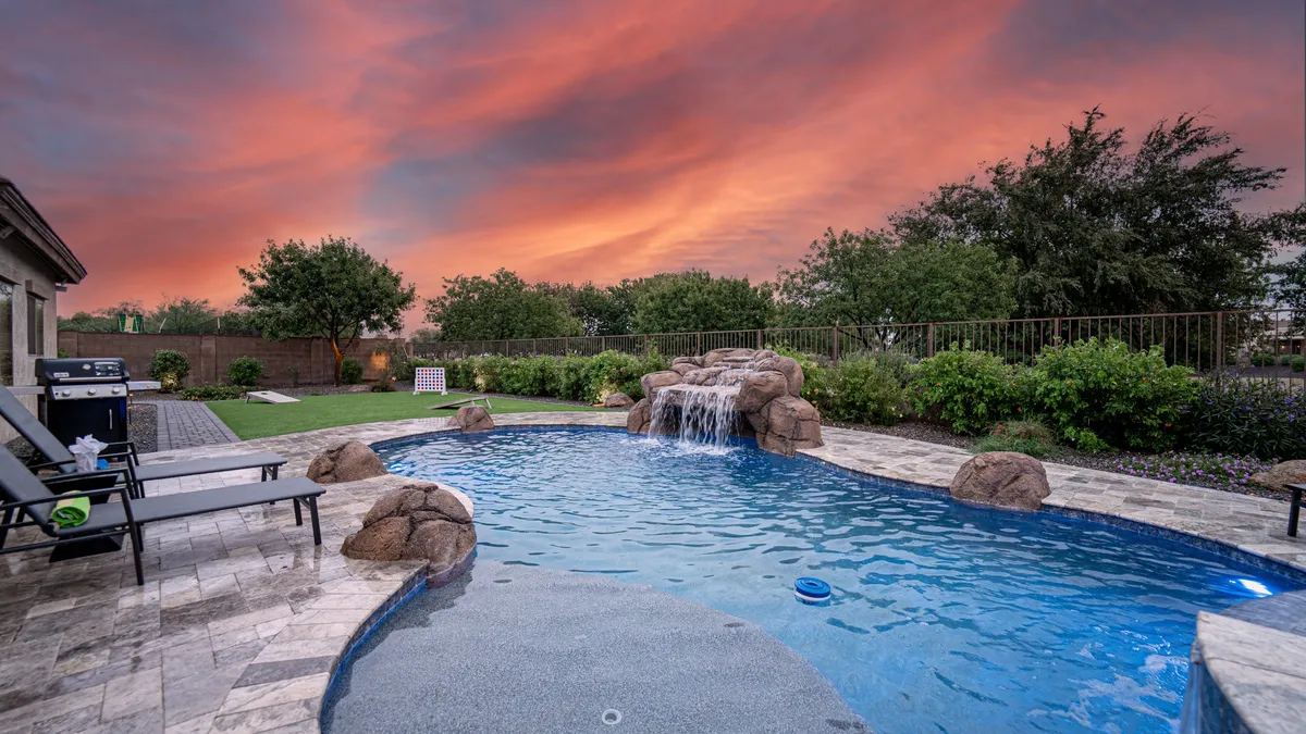 A serene outdoor scene featuring a sparkling swimming pool with a gentle waterfall, a sunset sky, and comfortable lounge chairs arranged for relaxation and socializing. The mood is tranquil, inviting guests to unwind or gather poolside.