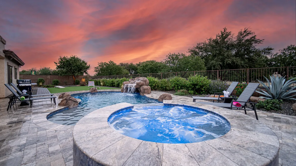 This image showcases a sparkling swimming pool with a spa hot tub, complemented by a soothing waterfall feature and surrounded by comfortable lounge chairs. The sky is tinged with sunset colors, giving the entire area a tranquil, luxurious atmosphere.
